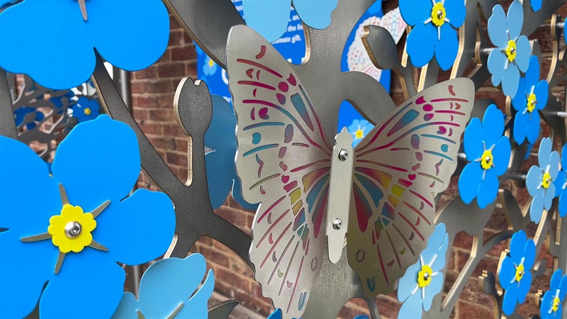 The installation includes intertwined shimmering foliage, vibrant butterflied, and forget-me-not flowers crafted from stainless steel